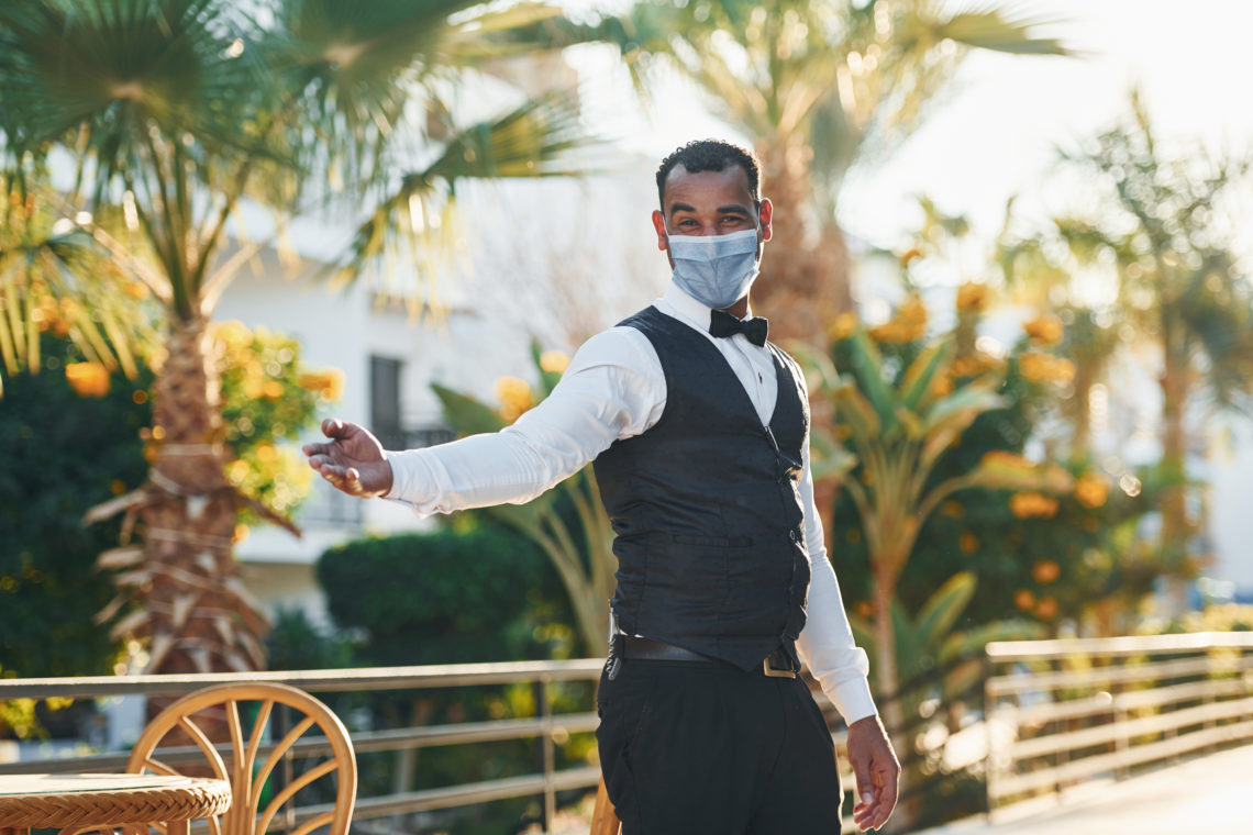 In protective mask. Black waiter in formal clothes is at his work outdoors at sunny daytime